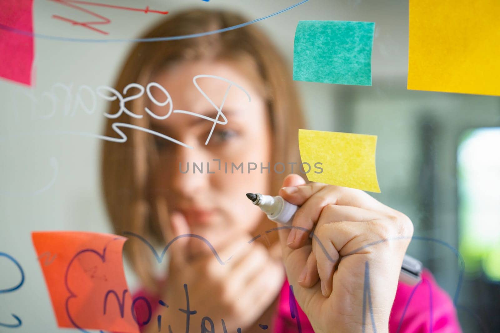 Closeup of young beautiful caucasian business leader presents marketing idea while writing marketing idea on glass board with mind map and colorful sticky notes. Portrait. Brainstorm. Immaculate.