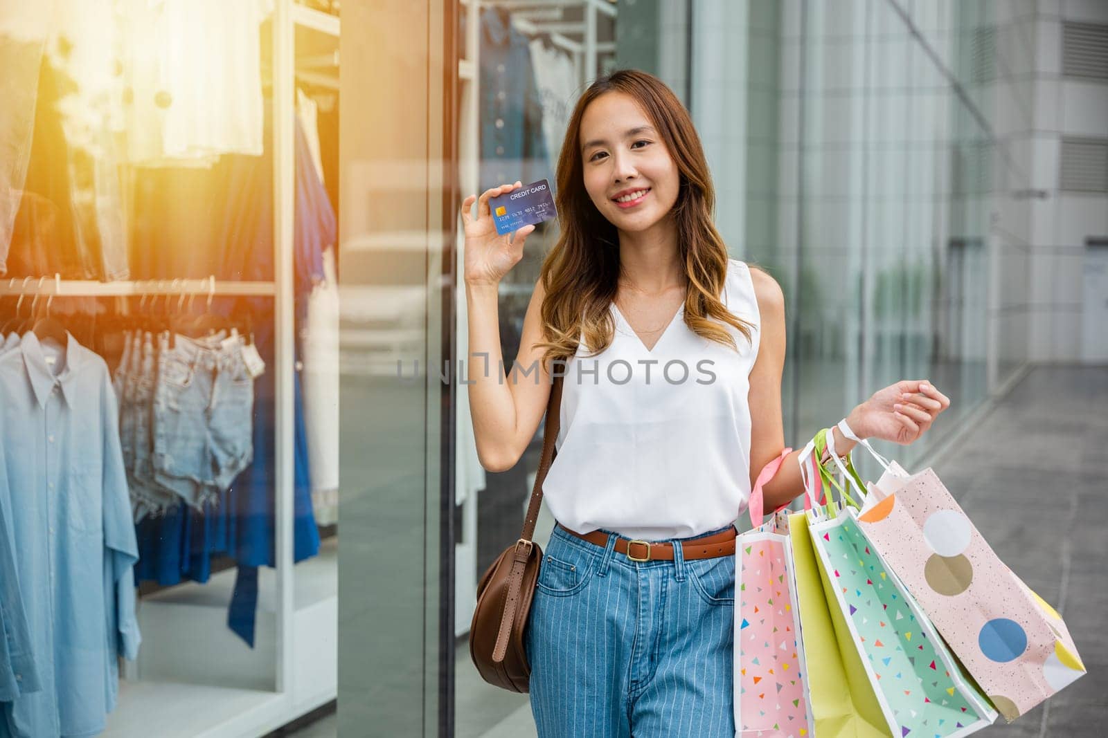 A young woman with a smile holds shopping bags and a credit card in front of a store window. She's ready to spend money and take advantage of the latest discounts and deals