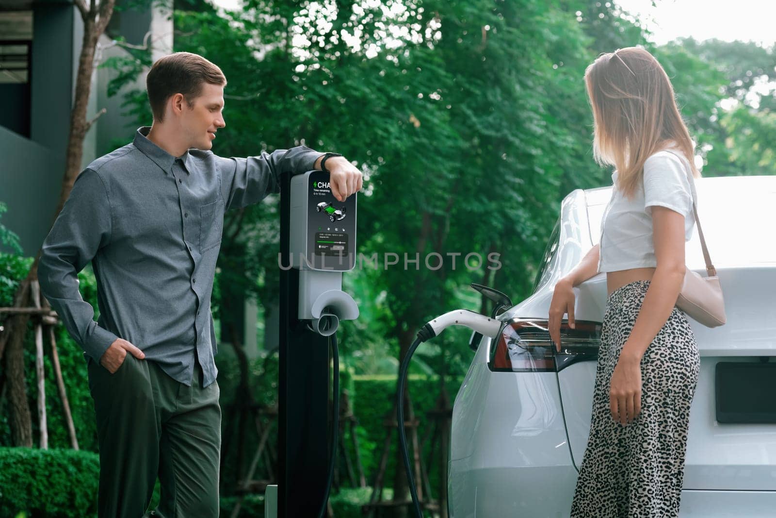 Young couple travel with EV electric car charging in green sustainable city outdoor garden in summer shows urban sustainability lifestyle by green clean rechargeable energy of electric vehicle innards