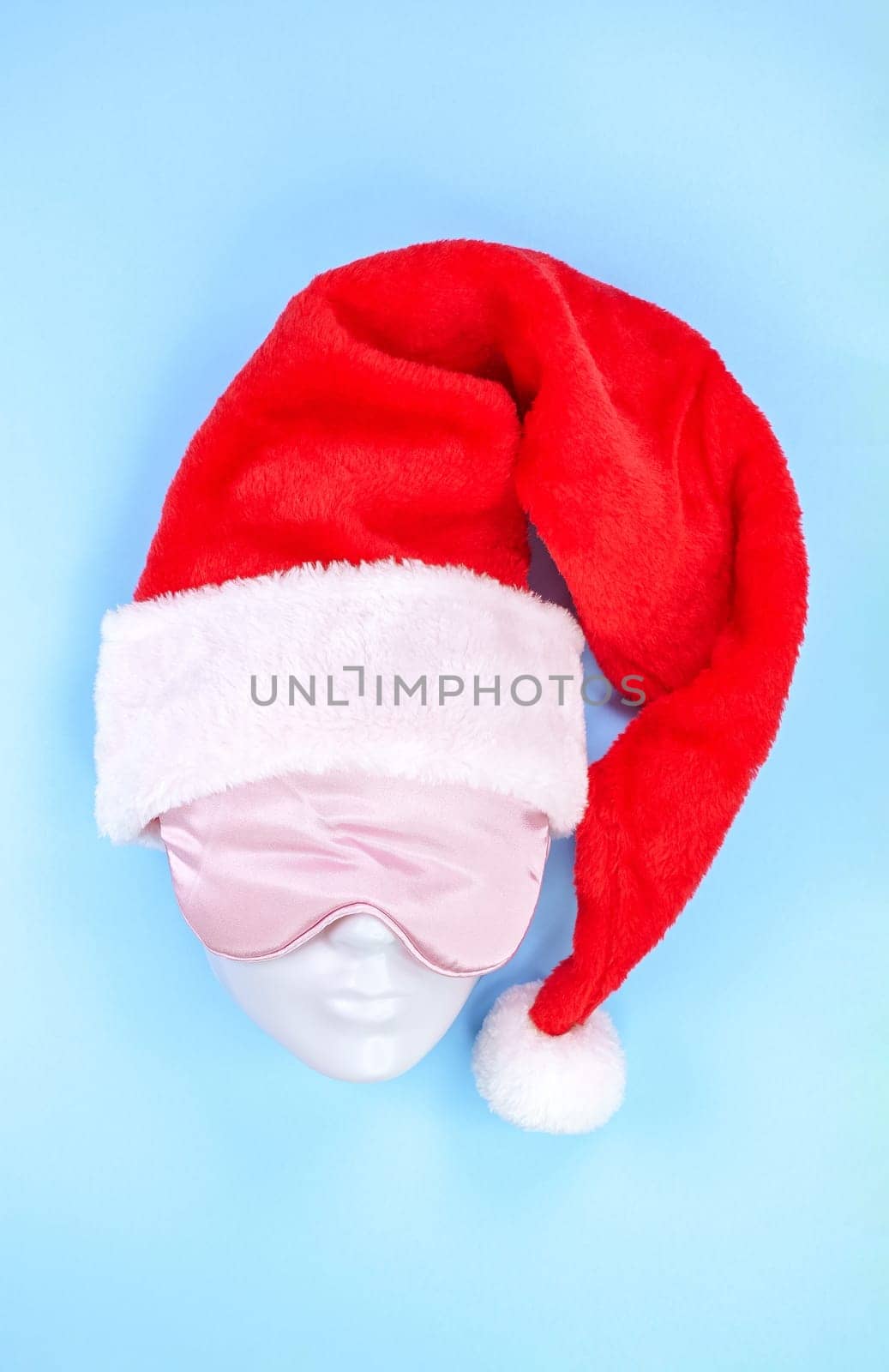 Pink sleeping eye mask on mannequin face with red christmas hat on blue background, sleeping disorder. Holidays, Head accessory. Last minute shopping sickness, Plastic face