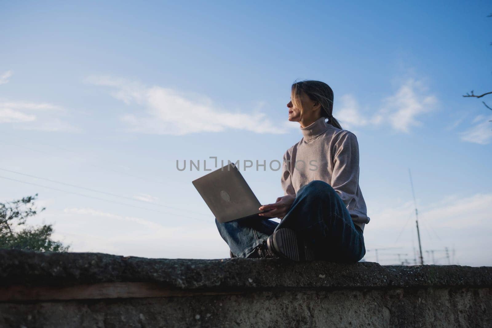 Woman freelancer uses laptop on cement wall outdoors against the sky. The woman to be focused on her work or enjoying some leisure time while using her laptop. by Matiunina