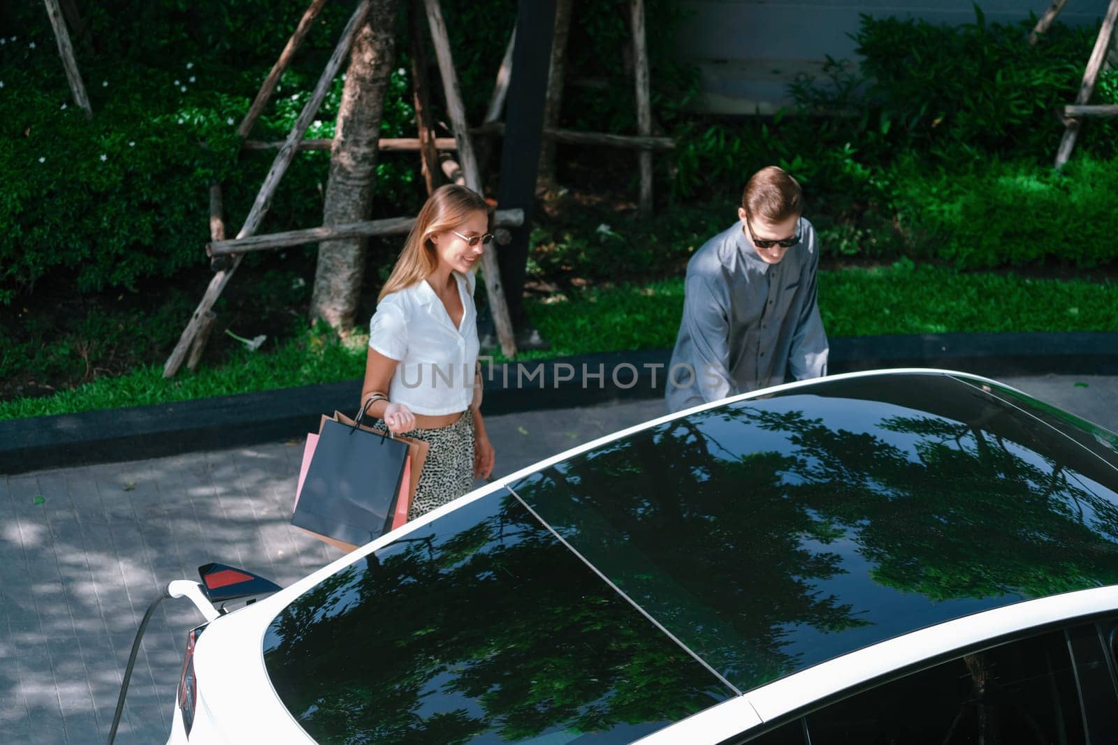 Young couple travel with EV electric car charging in green sustainable city outdoor garden in summer shows urban sustainability lifestyle by green clean rechargeable energy of electric vehicle innards