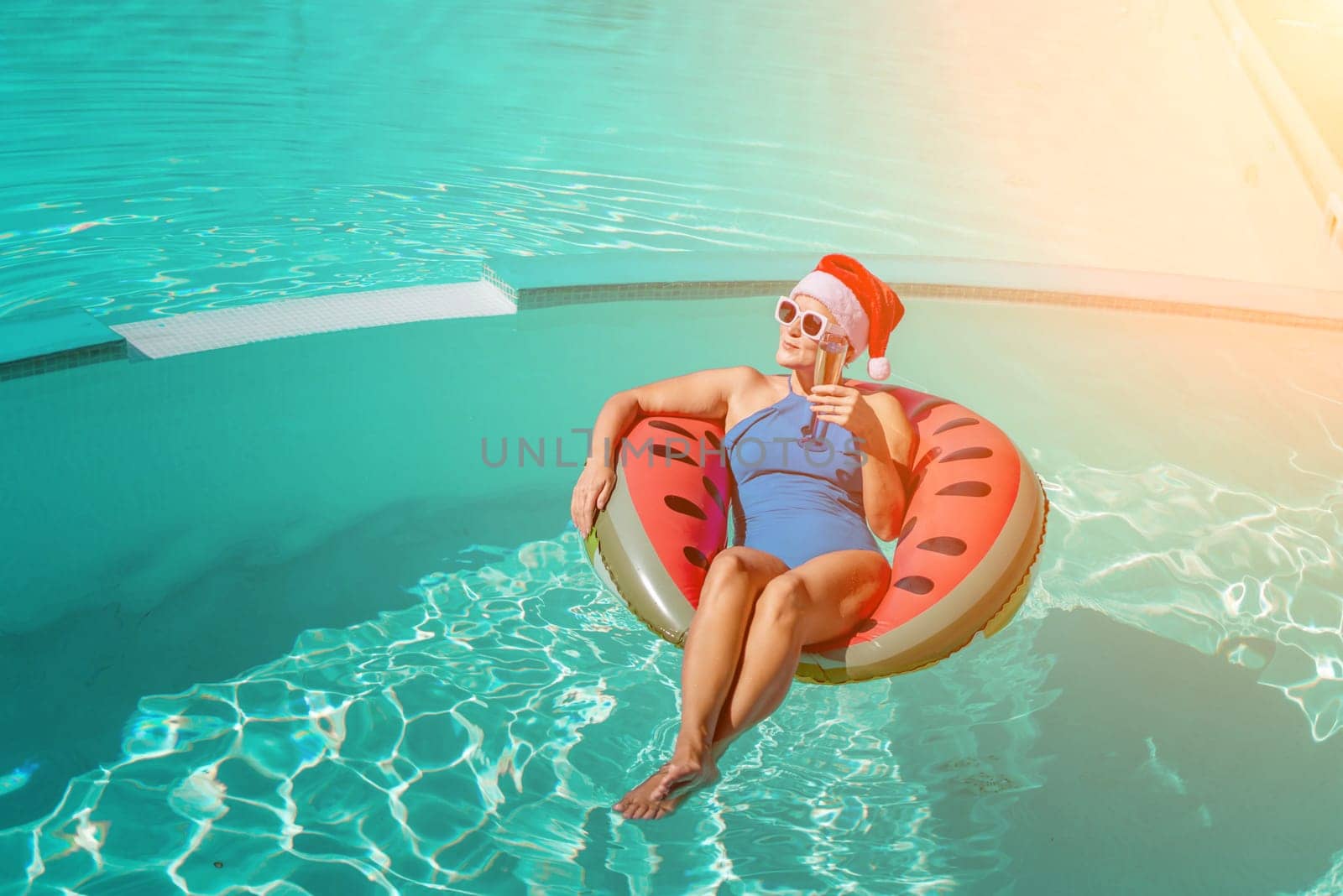 A happy woman in a blue bikini, a red and white Santa hat and sunglasses poses in the pool in an inflatable circle with a watermelon pattern, holding a glass of champagne in her hands. Christmas holidays concept. by Matiunina