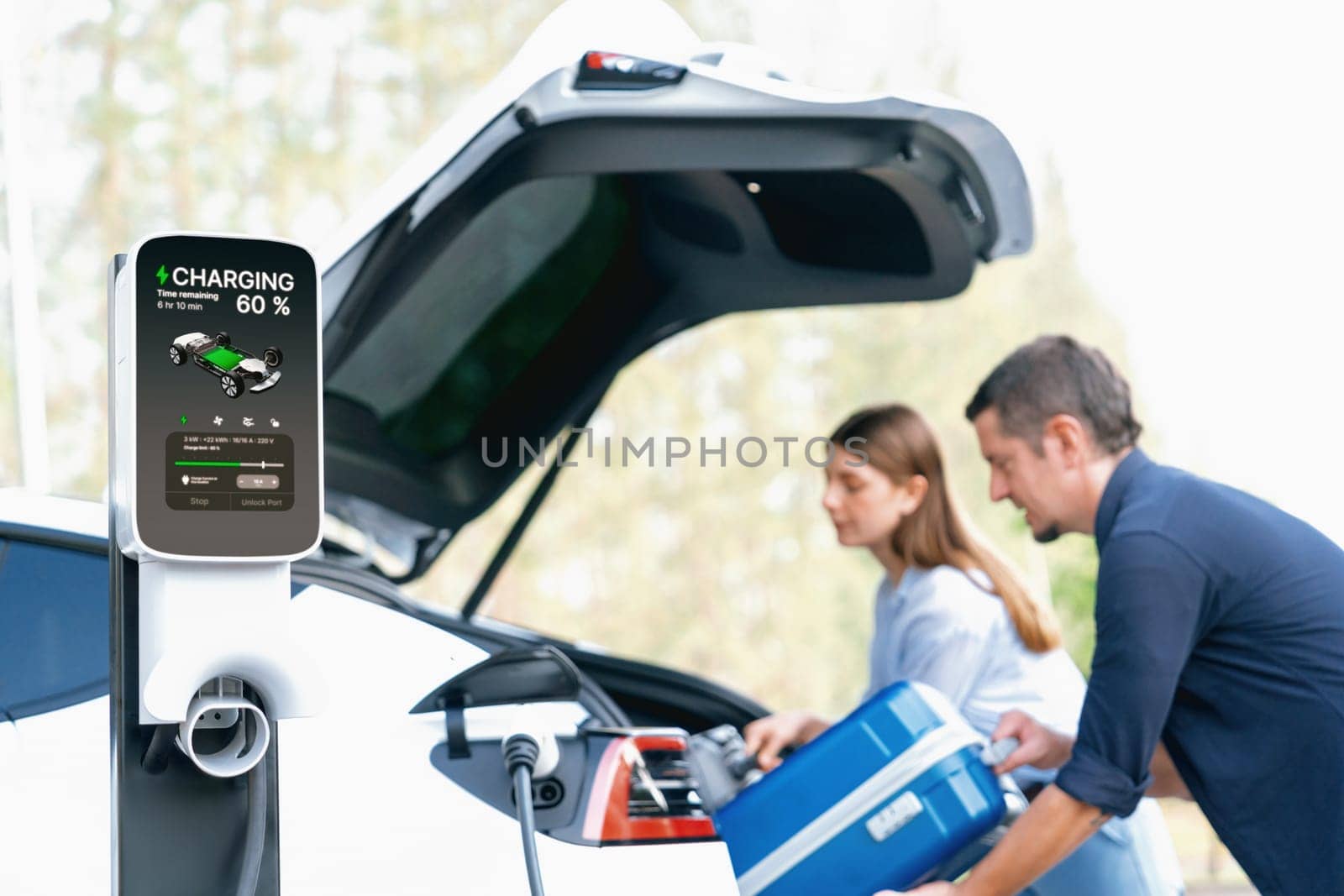 Lovely young couple recharging battery for electric car while relaxing and sitting on the trunk during road trip travel EV car in autumnal forest. Eco friendly travel on vacation during autumn. Exalt