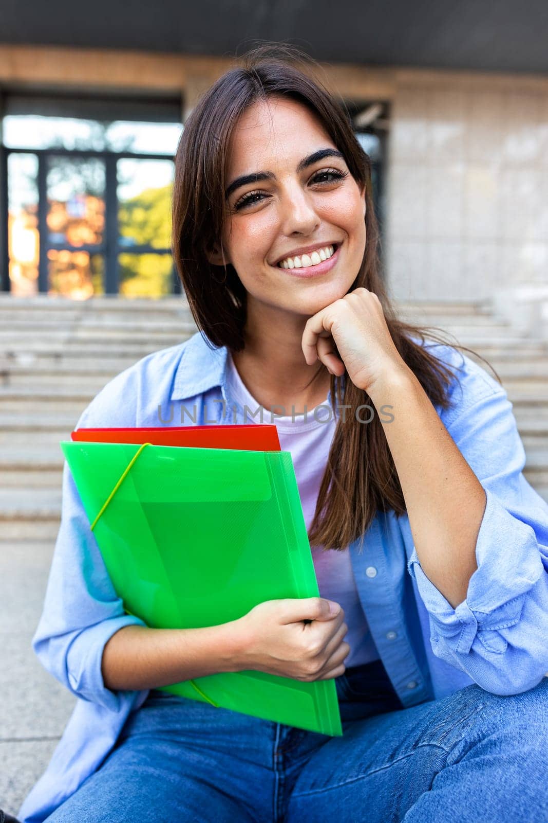 Vertical portrait of young beautiful female college student sitting outside university building looking at camera. Education concept.