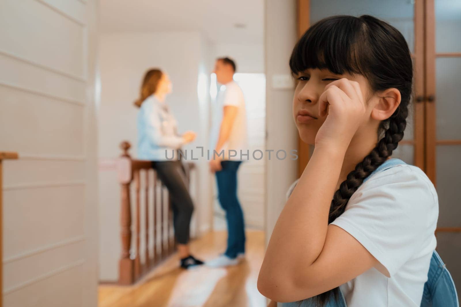 Stressed and unhappy young girl huddle in corner wiping her tears while her parent arguing in background. Domestic violence at home and traumatic childhood develop to depression. Synchronos