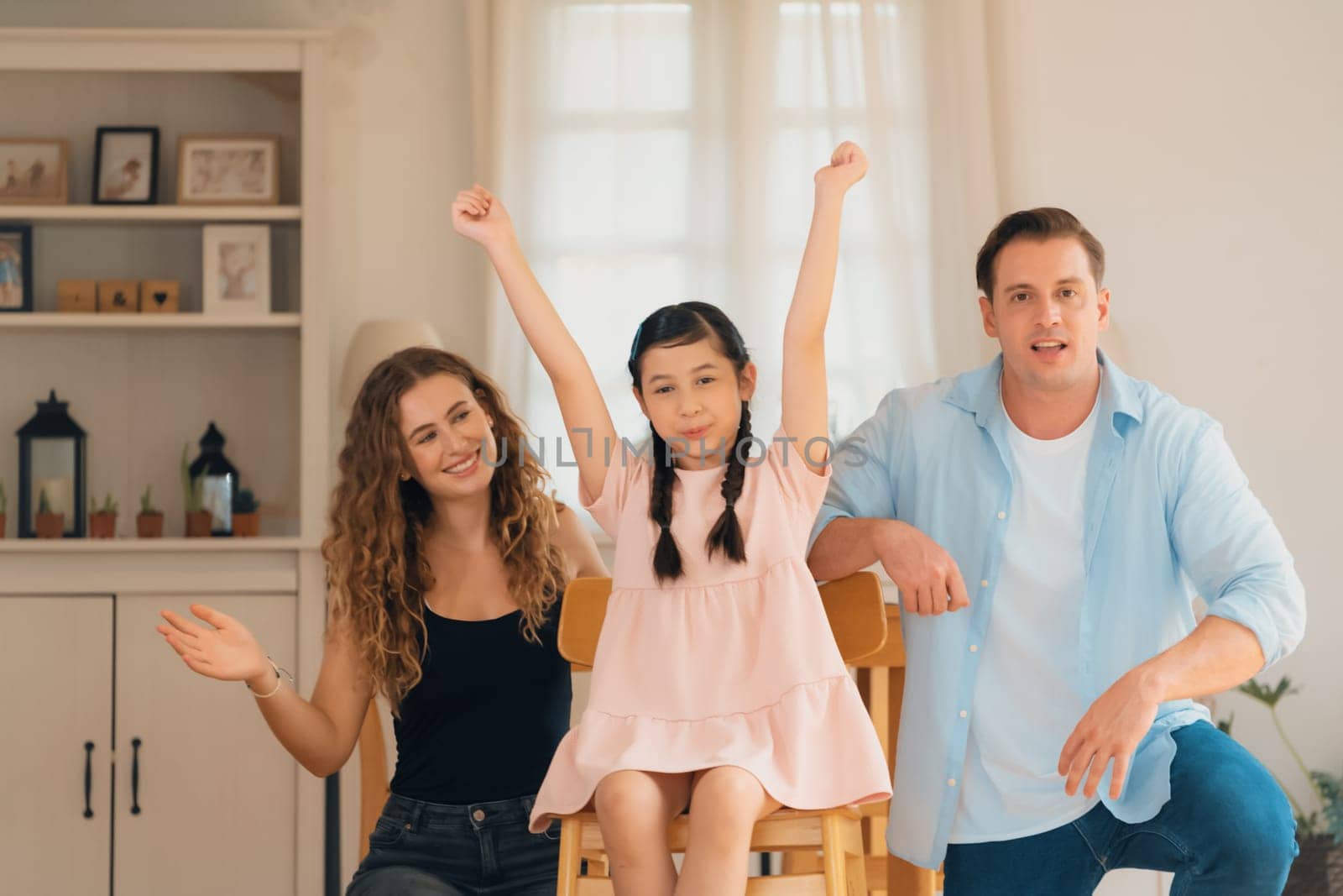 Happy family portrait with lovely little girl smiling and looking at camera, lovely and cheerful parent and their daughter sitting together in living room at home with warm daylight. Synchronos