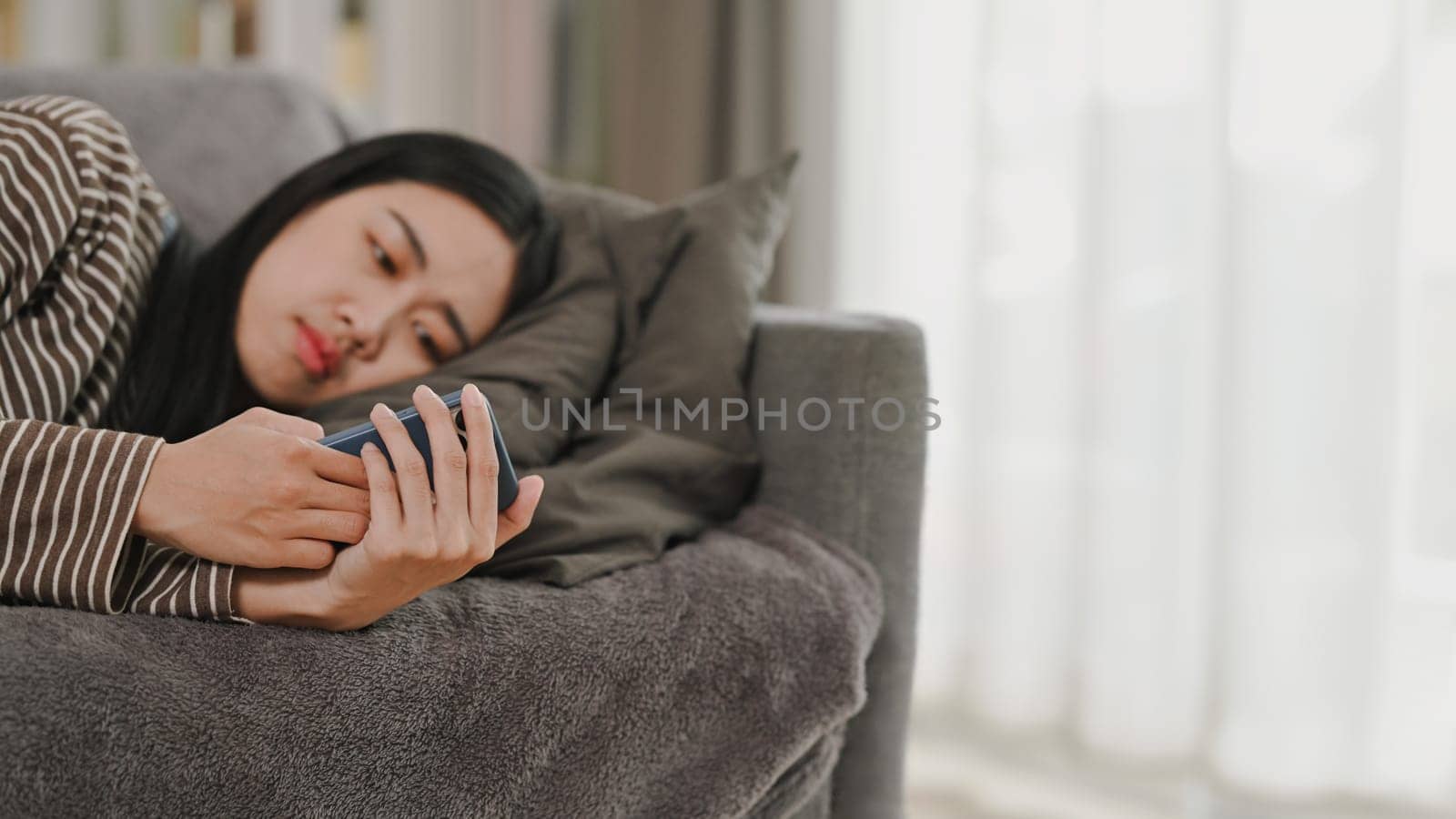 Bored young woman in casual clothes using mobile phone while lying on sofa at home. People lifestyle concept.