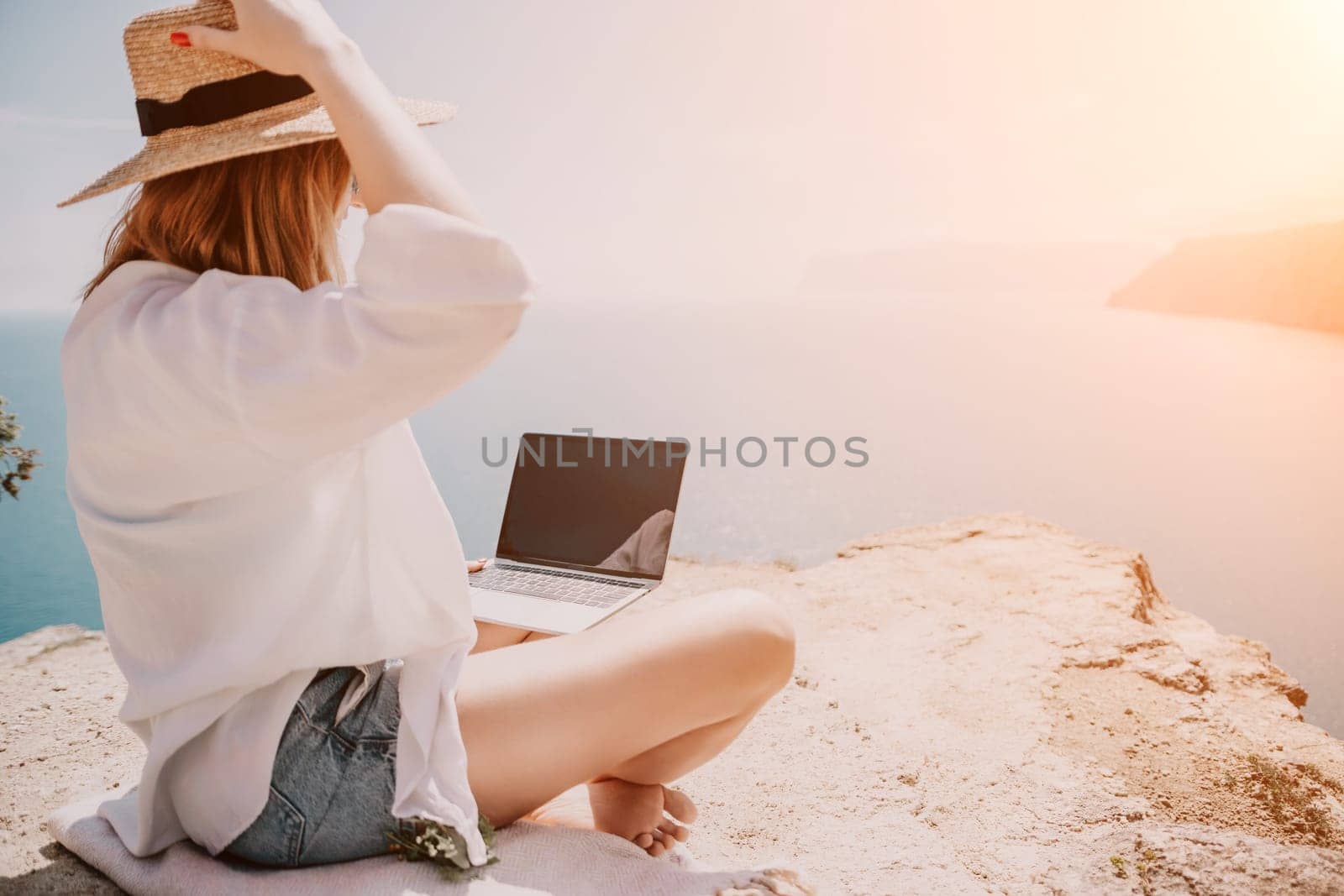 Successful business woman in yellow hat working on laptop by the sea. Pretty lady typing on computer at summer day outdoors. Freelance, travel and holidays concept.