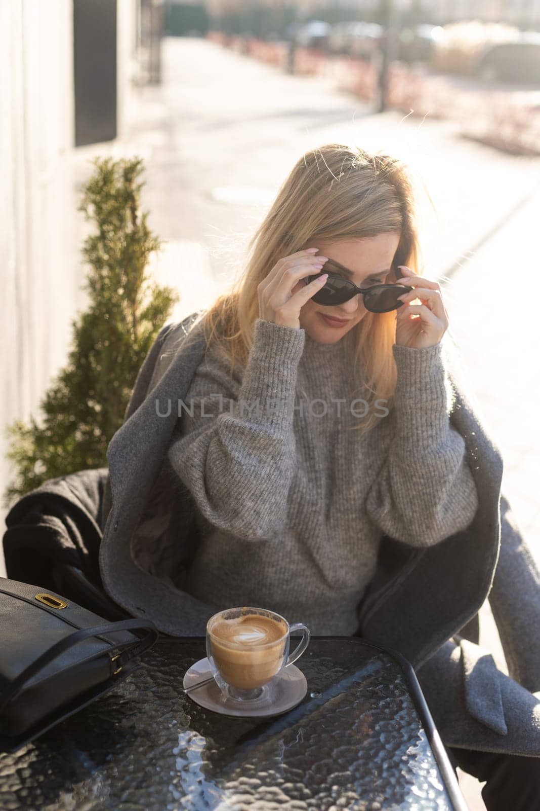 Young woman enjoying a coffee, sitting with mobile phone on the cafe terrace on the old city street during a sunny day by Andelov13