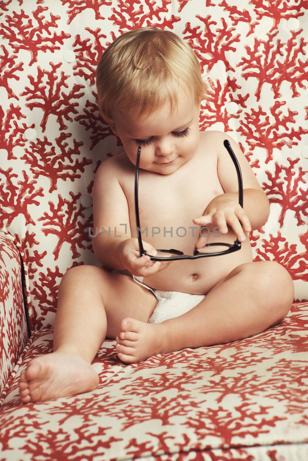 Happy baby, blonde boy and playing with glasses on sofa in growth, youth or development in living room at home. Cute, adorable or little toddler, kid or child smile with innocent or positive attitude.