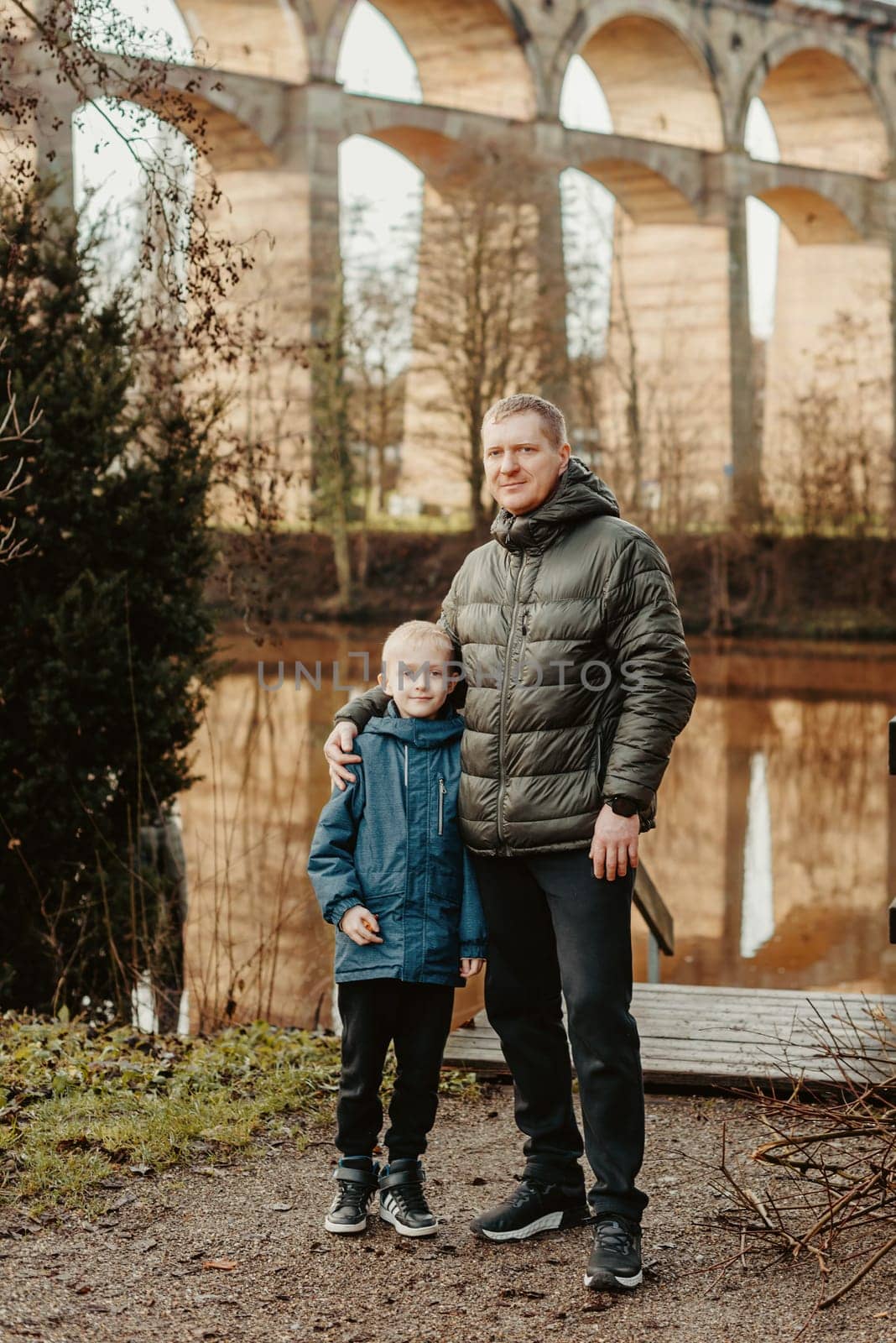 Family Serenity: Handsome 40-Year-Old Man and 8-Year-Old Son Amidst the Beauty of Neckar River and Historic Bridge, Bietigheim-Bissingen, Germany, Winter or Autumn. Embrace the warmth of family bonds against the backdrop of scenic beauty. This image features a handsome 40-year-old man and his 8-year-old son standing in the serene park by Neckar River and Historic Bridge in Bietigheim-Bissingen, Germany. Whether touched by winter's chill or the colors of autumn, it's a timeless moment of familial connection amidst captivating surroundings.