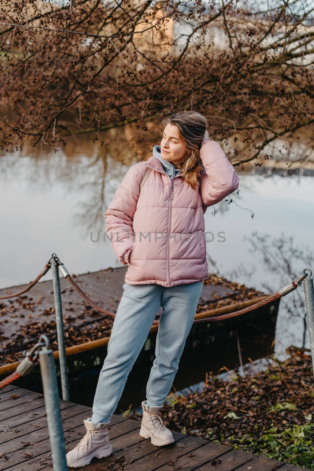 A young woman standing at the shore looking at the river in autumn sunny day. Street view, copy space for text, travel photo. Happy tourist woman on the bank of the river in autumn in warm clothes. Tourists enjoy their vacation, winter season. Romantic look and travel concept. A joyful mood in a Caucasian girl. Winter Wonderland: Enchanting Girl by the Riverside in Autumn.