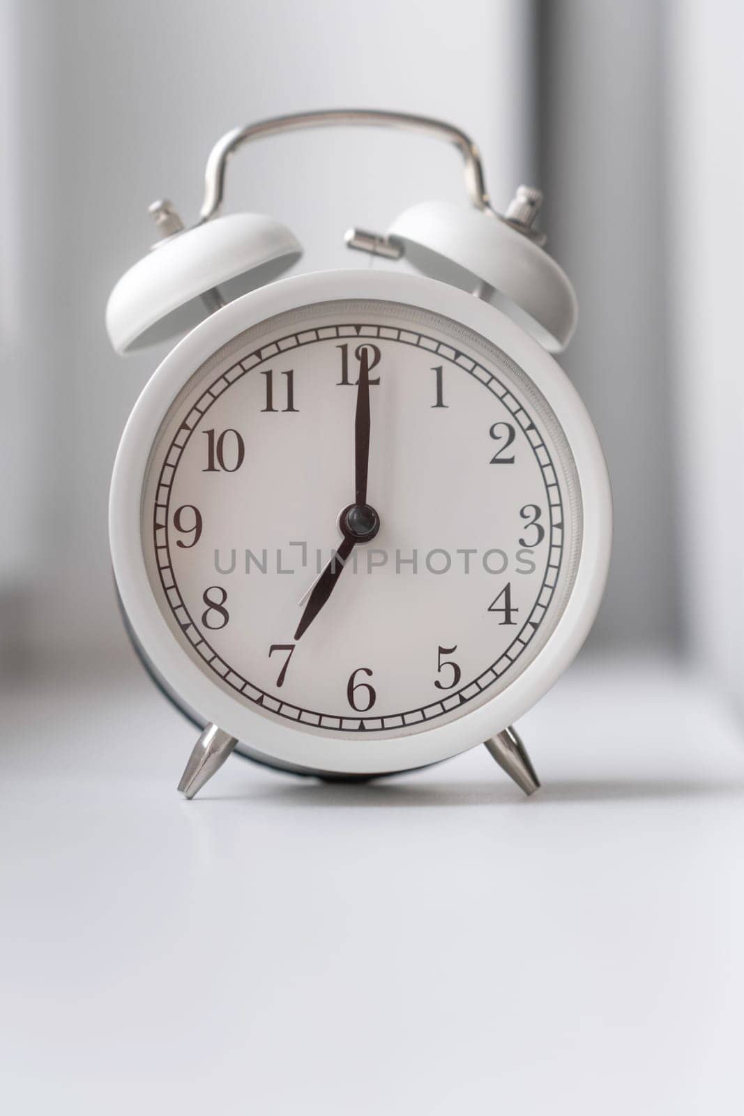 Close-up of a round white alarm clock on the windowsill in the bedroom. The hands on the clock show seven o'clock in the morning, time to get up. Retro alarm clock on the table. space for text