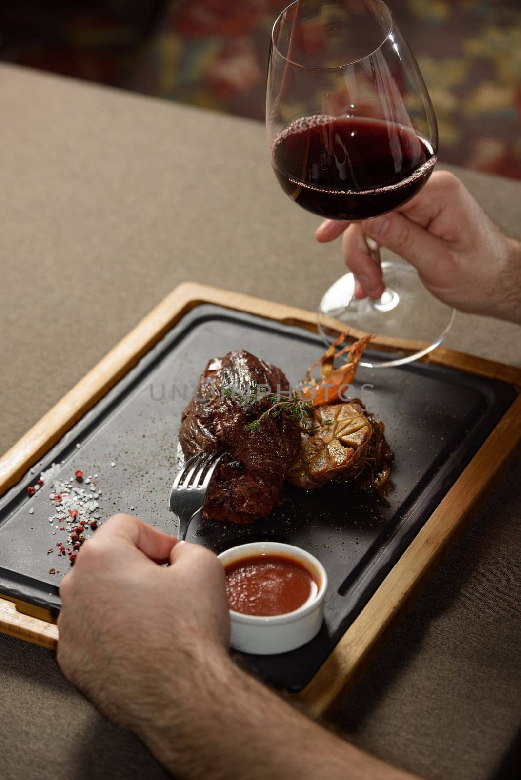 man sitting at a table in a restaurant. He is holding a glass of red wine and eating a steak.