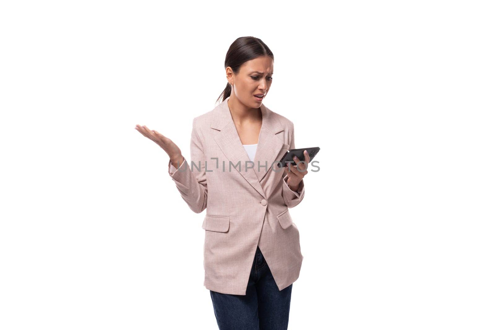 young confident caucasian woman with a ponytail hairstyle dressed in a jacket and jeans uses a smartphone in her hands by TRMK