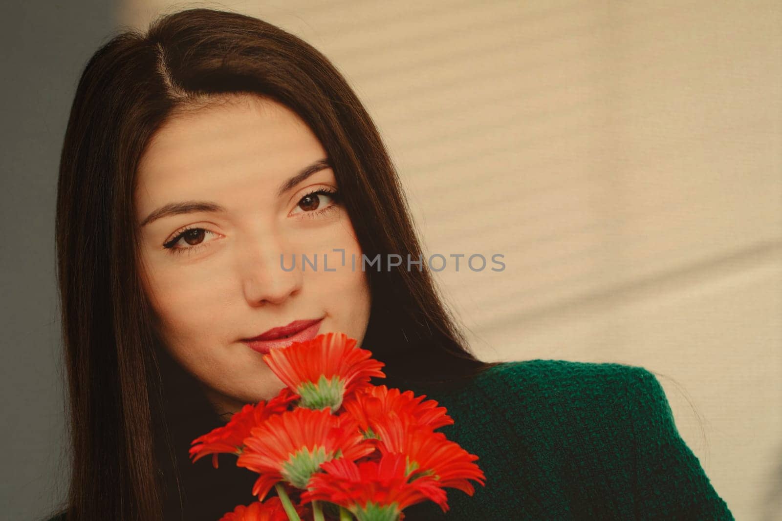 Portrait of one beautiful young Caucasian brunette girl with brown eyes, long hair in a green fashionable jacket holding a bouquet of red gerbernas near her face against a white wall, close-up side view.