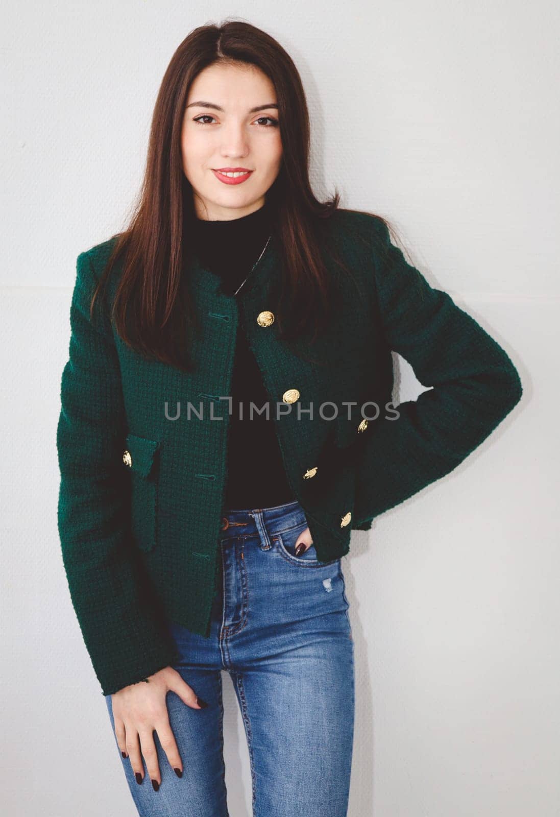 Portrait of one beautiful young Caucasian brunette with brown eyes, long and straight hair in a green fashionable jacket, blue jeans standing with a smile on her face against the background of a white wall in the room, close-up side view.