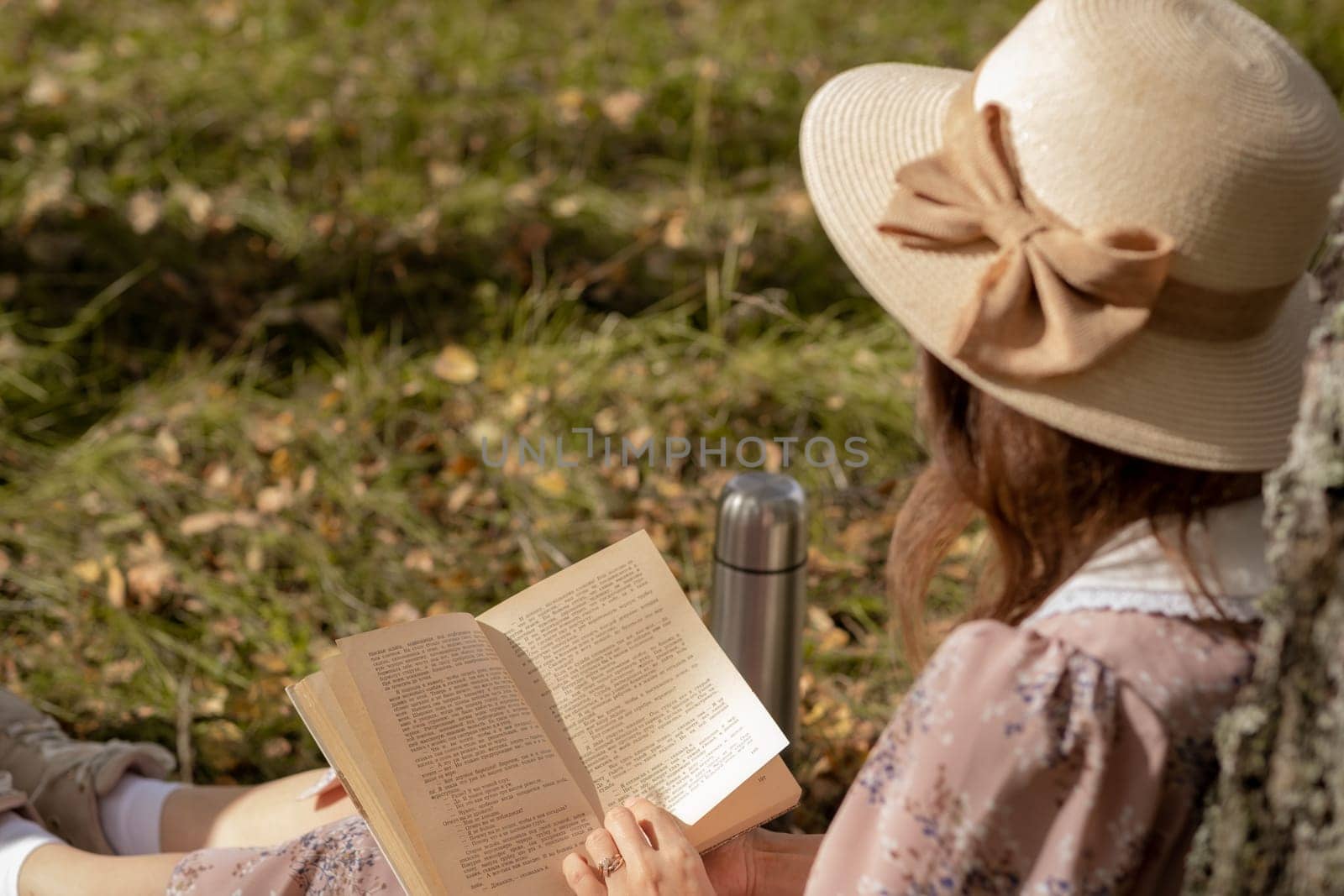 A young beautiful woman in a dress and a round hat reads a book outdoors in the forest and drinks tea. Romantic and vintage photo of a beautiful girl. Reading and relaxation