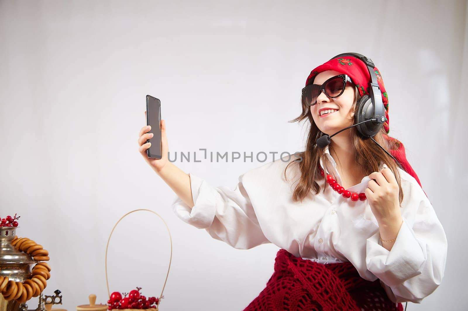 A fashionable modern girl in stylized folk clothes at a table with a samovar, bagels and tea takes a selfie on the Orthodox holiday of Maslenitsa and Easter. Funny photo shoot for a young woman by keleny