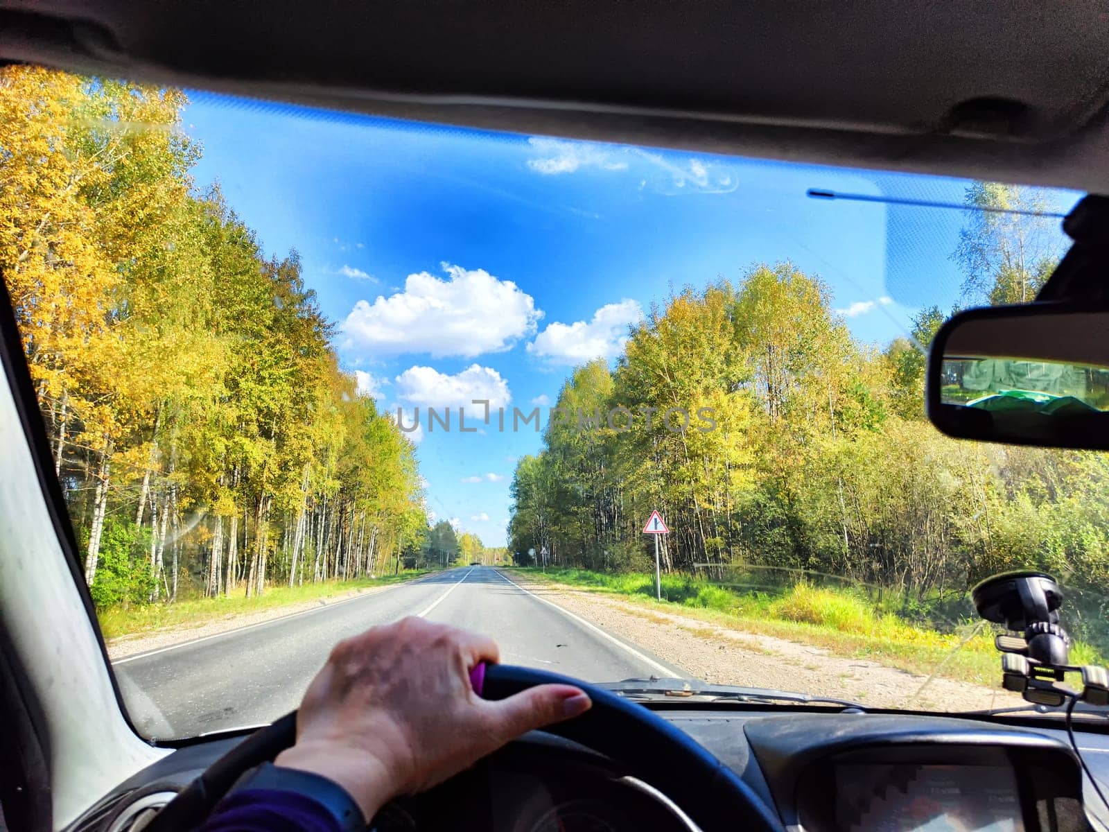 view from a car windshield of natural landscape with road, green trees and blue sky in summer or spring time. Hand of woman on steering wheel. Female traveler driving alone on trip or journey