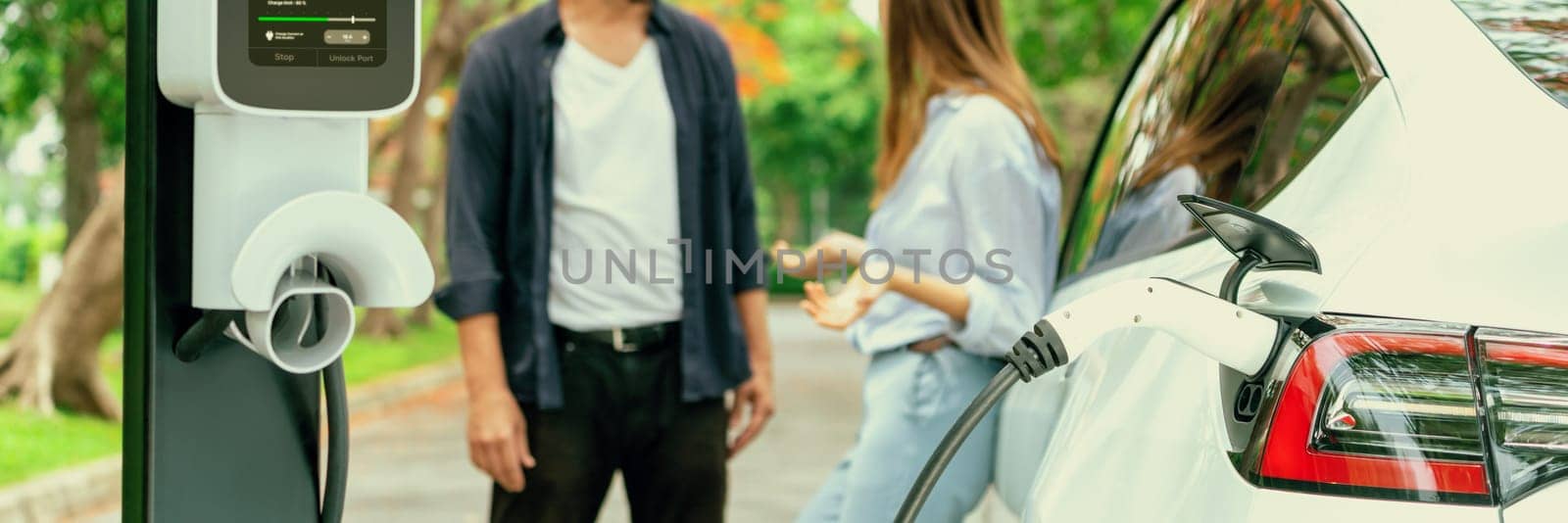 Panorama banner of lovely young couple recharging battery for electric car during autumnal road trip travel EV car in autumnal forest. Eco friendly travel on vacation during autumn. Exalt