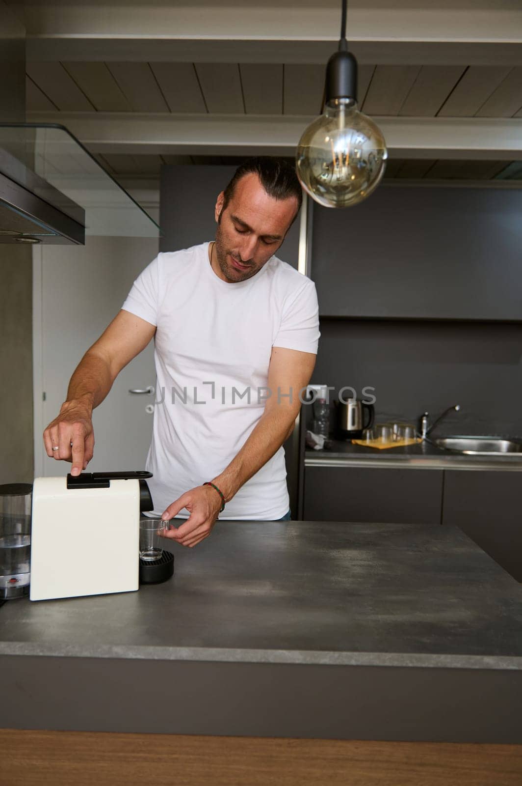 Caucasian handsome young man inserting a capsule with grinded coffee inside a coffee machine for home use, preparing freshly brewed espresso for breakfast in the morning, standing at kitchen counter