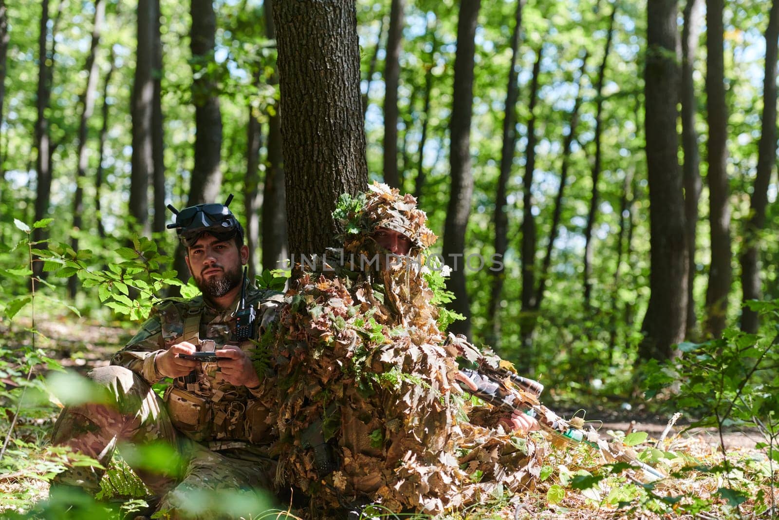 A skilled sniper and a soldier operating a drone with VR goggles strategize and observe the military action while concealed in the forest.