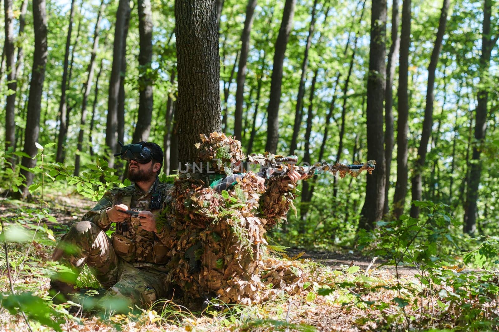 A skilled sniper and a soldier operating a drone with VR goggles strategize and observe the military action while concealed in the forest.