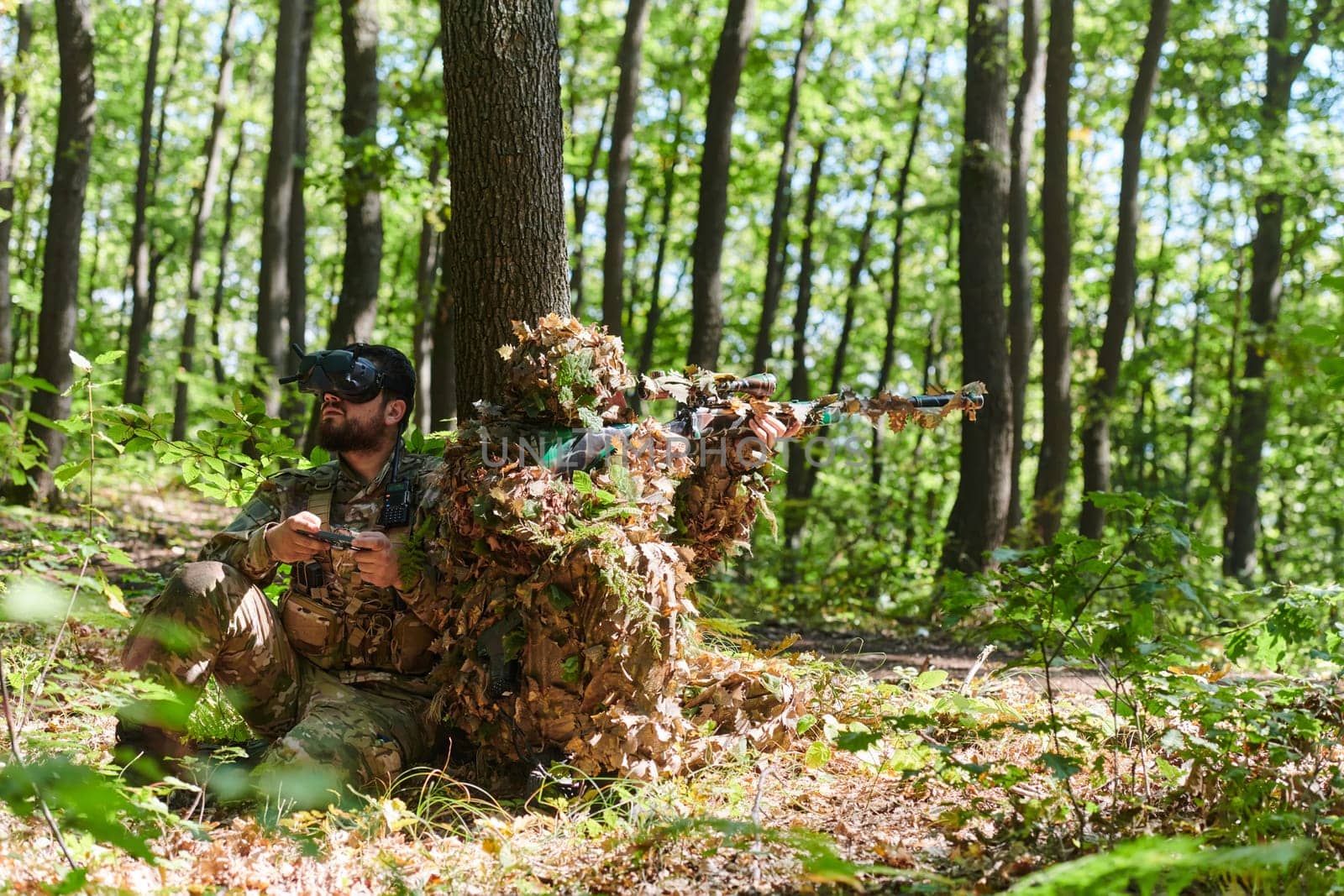 A skilled sniper and a soldier operating a drone with VR goggles strategize and observe the military action while concealed in the forest.