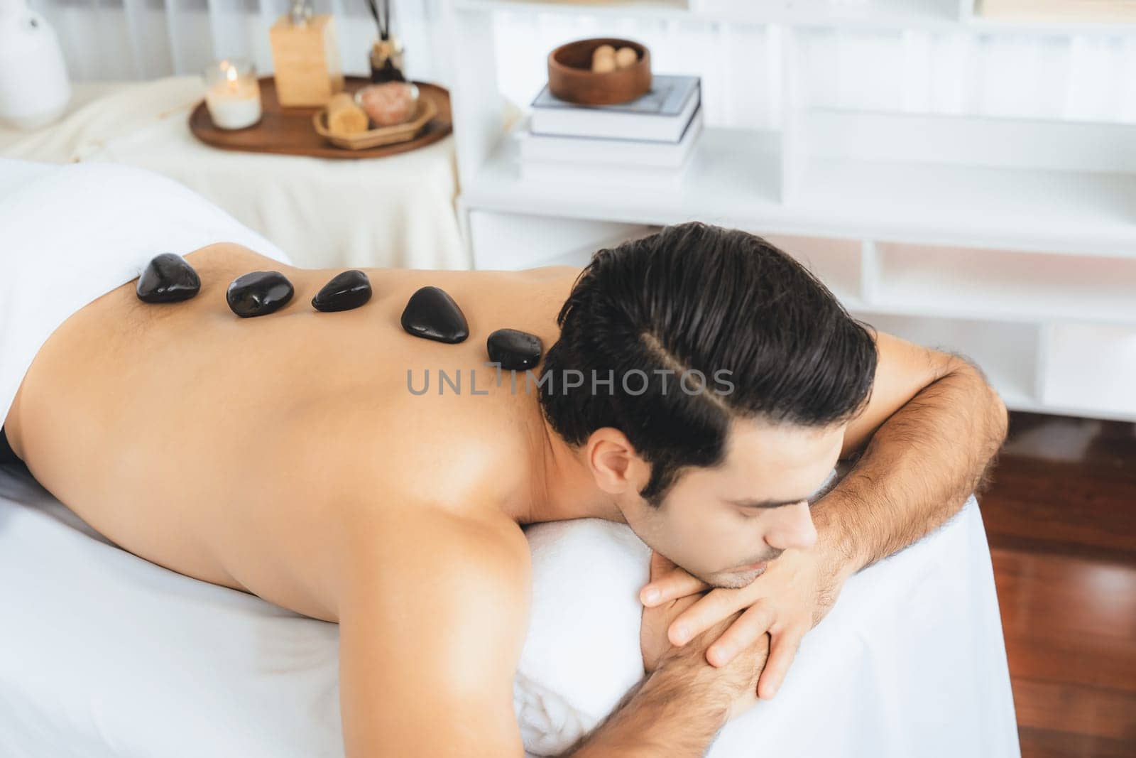 Hot stone massage at spa salon in luxury resort with day light serenity ambient, blissful man customer enjoying spa basalt stone massage glide over body with soothing warmth. Quiescent