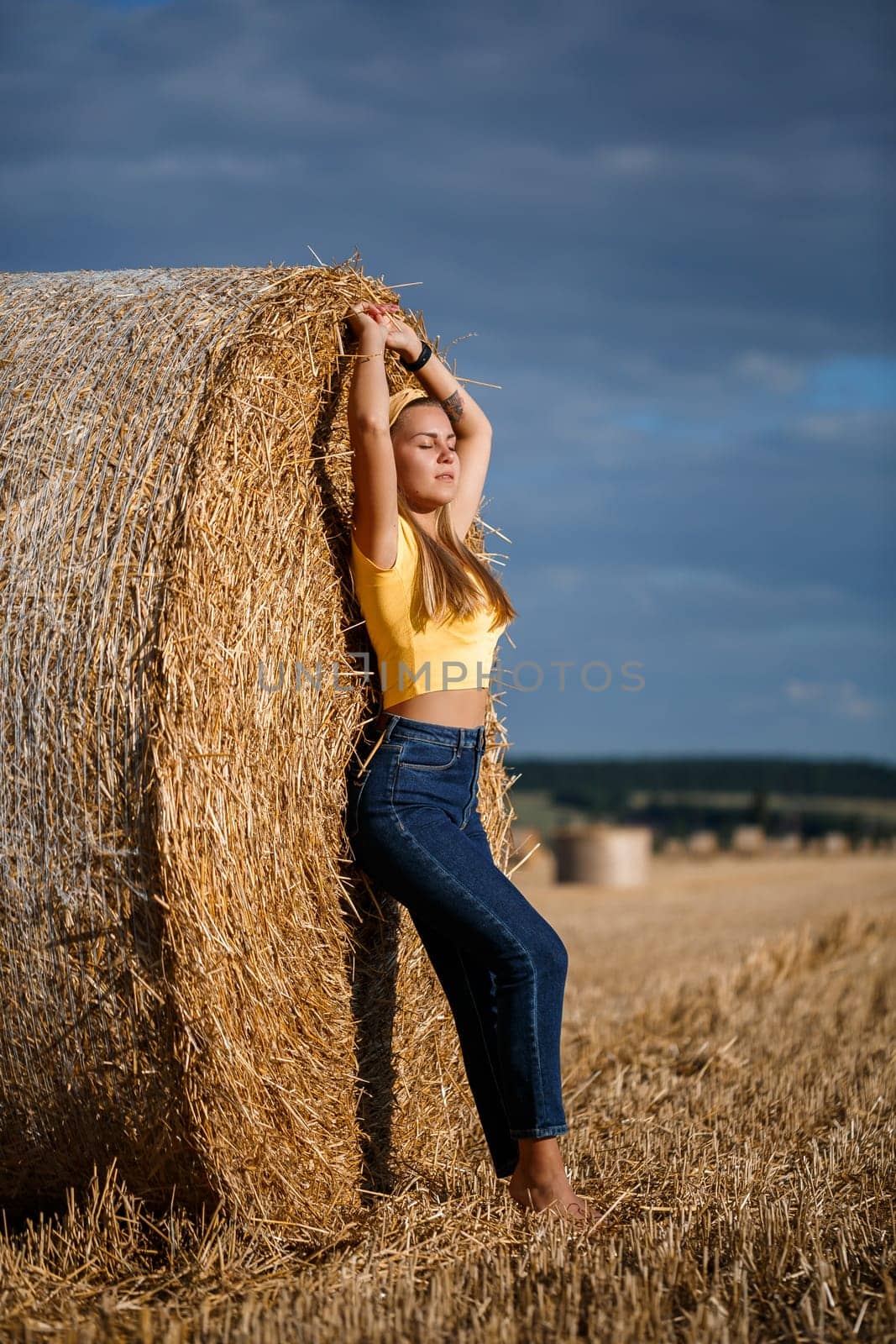 A young beautiful blonde stands on a mown wheat field near a huge sheaf of hay, enjoying nature. Nature in the village