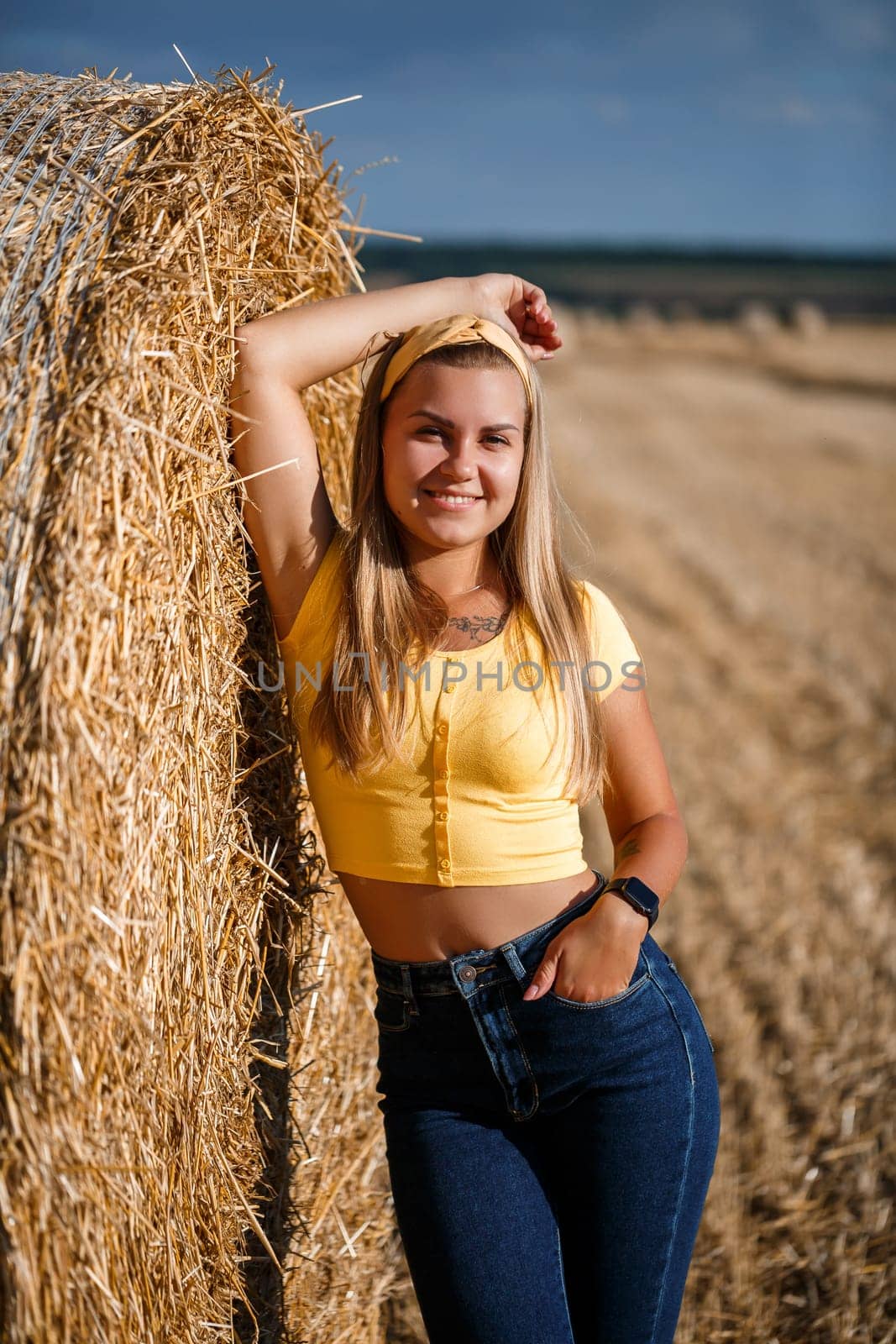 A young beautiful blonde stands on a mown wheat field near a huge sheaf of hay, enjoying nature. Nature in the village