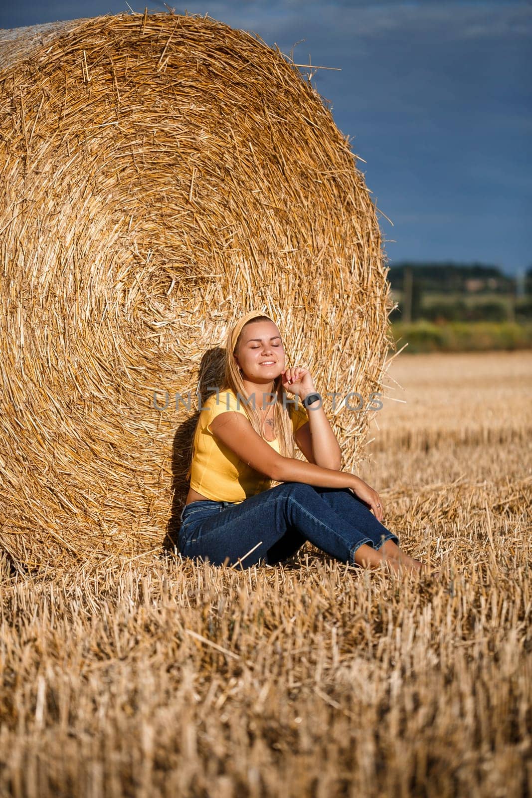 Beautiful young woman near a sheaf of hay in a field. Holidays in the village, a girl enjoying nature in a mown field on a sunny day