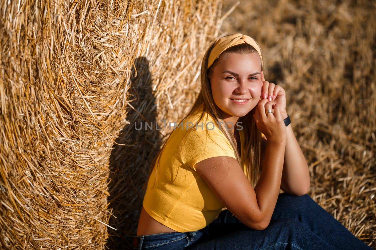 A young beautiful girl in a field stands near a sheaf. Vacation in the village. Young woman in yellow top and jeans