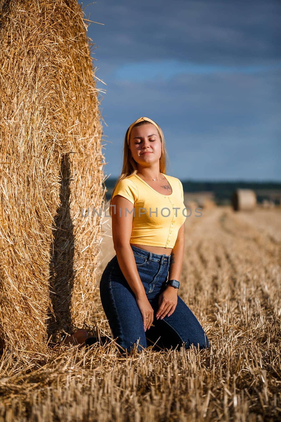 A young beautiful blonde stands on a mown wheat field near a huge sheaf of hay, enjoying nature. Nature in the village