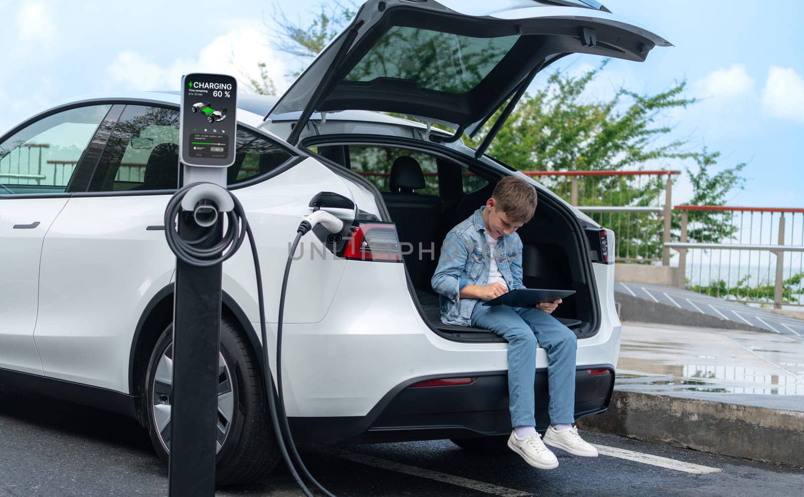 Little boy sitting on car trunk, using tablet while recharging eco-friendly car from EV charging station. EV car road trip travel as alternative vehicle using sustainable energy concept. Perpetual