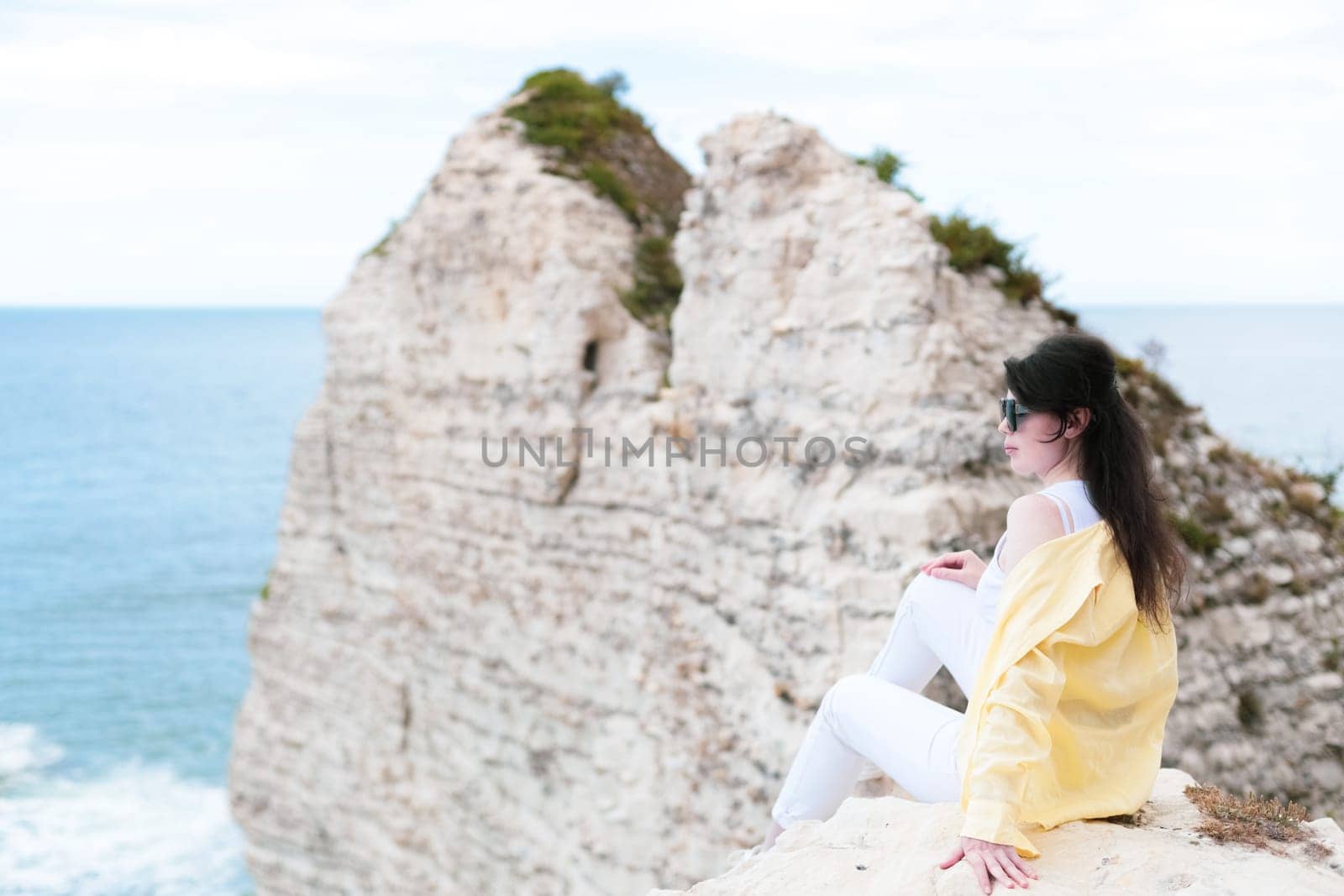 One young beautiful Caucasian brunette girl in white pants, a yellow shirt and sunglasses sits half-side high on a rock admiring the north sea in Etretat, Normandy, France, close-up view from below.
