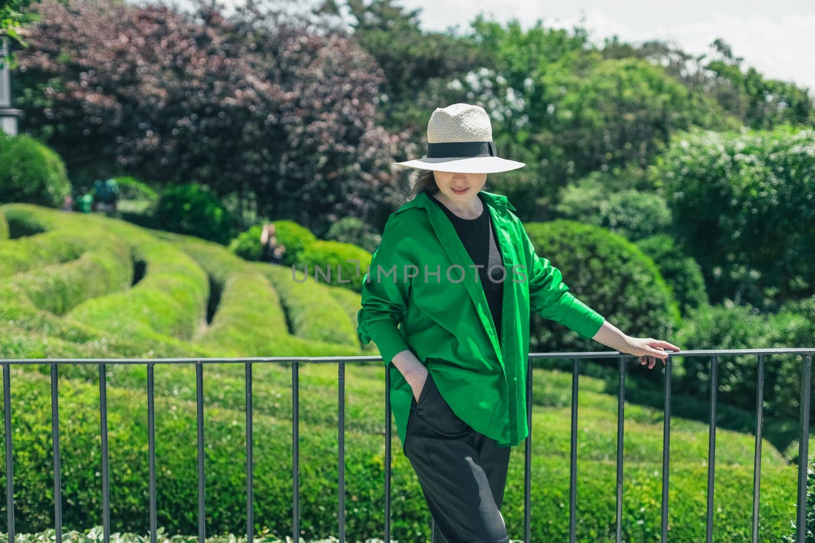 A portrait of one beautiful Caucasian young brunette girl in a straw hat pulled halfway down her face and a green shirt stands holding the railing in the botanical garden of wonders on a sunny summer day in France, side view close-up with depth of field.