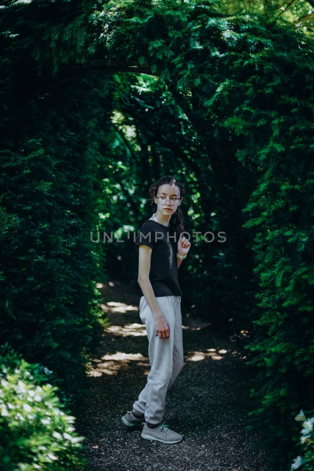 Portrait of one beautiful Caucasian young teenage girl in glasses and sportswear standing on a path with a plant arch in the botanical garden of wonders on a summer sunny day in France, close-up side view with depth of field.