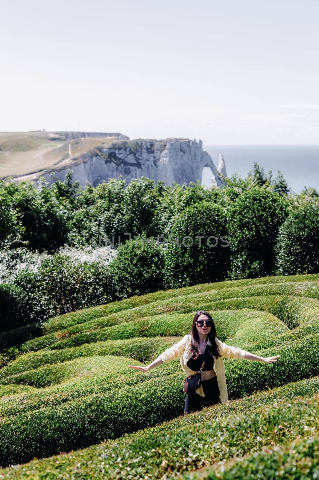 Portrait of one beautiful Caucasian young girl in sunglasses and a yellow shirt standing in spiral bushes with her arms spread apart in the Botanical Garden of Wonders on a summer sunny day in France, close-up side view with depth of field.