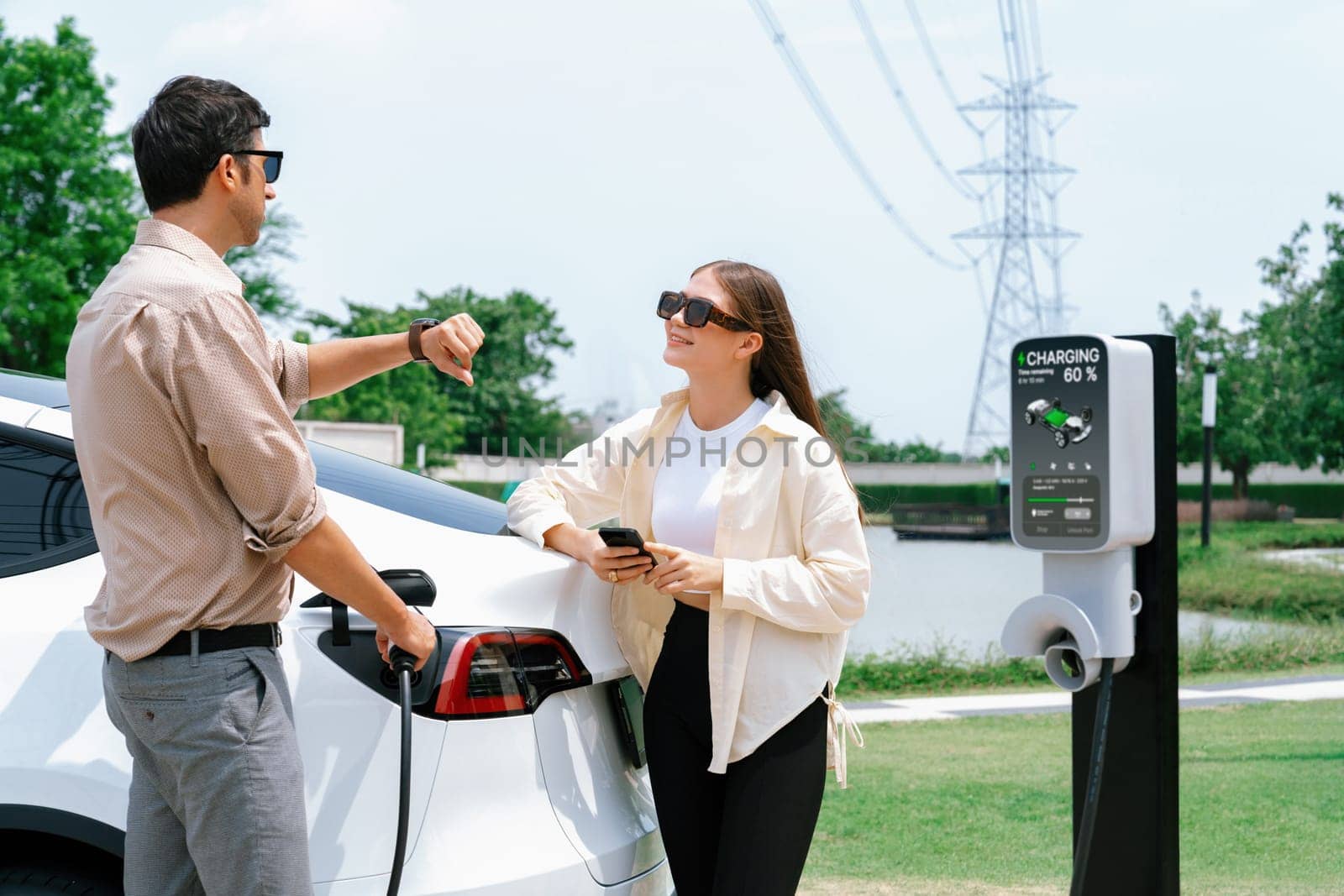 Young couple use smartphone to pay for electricity at public EV car charging station green city park. Modern environmental and sustainable urban lifestyle with EV vehicle. Expedient