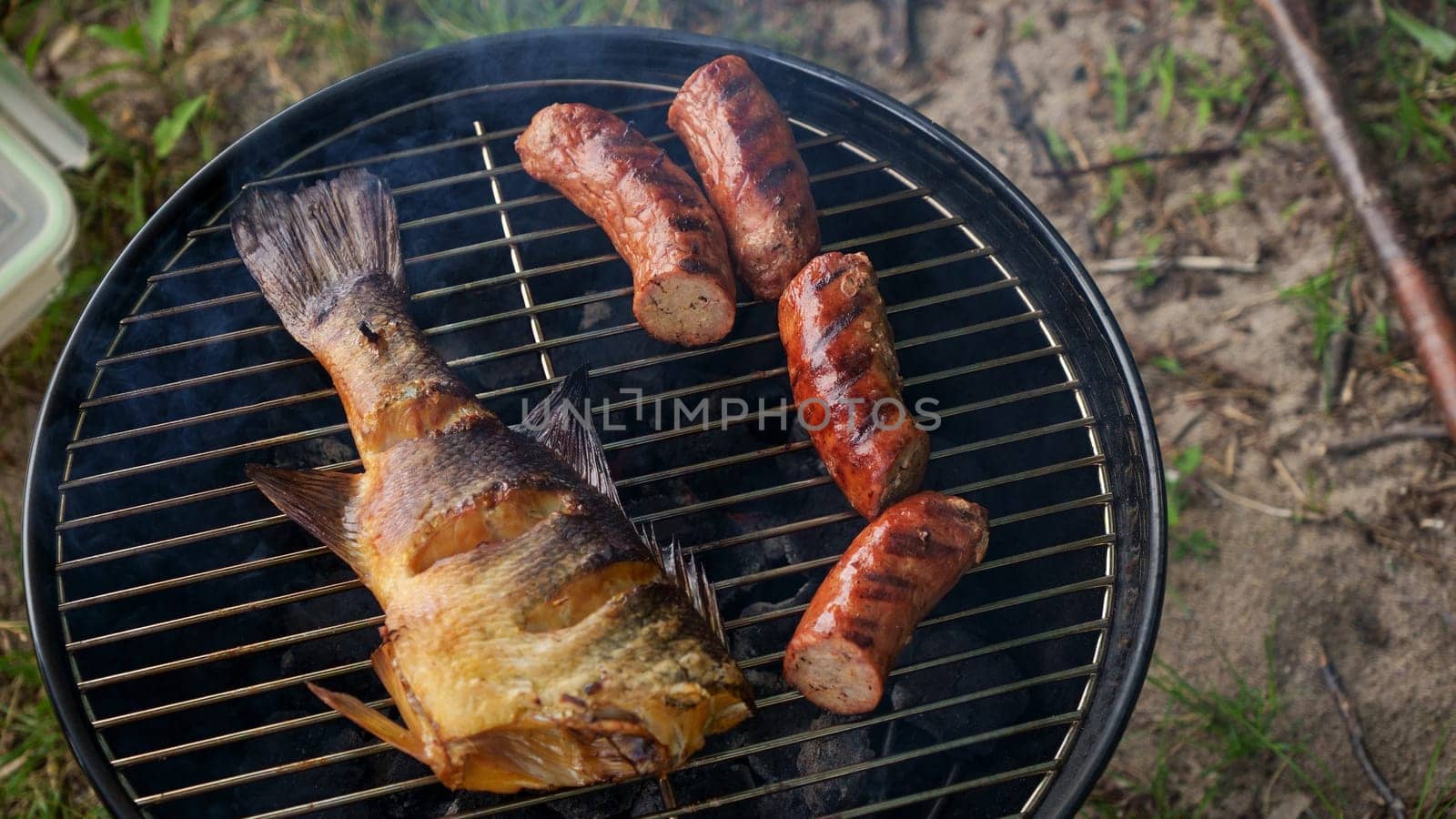Top view of fresh grilled whole fish, skewer, sausages on round black charcoal grill, green grass background. Barbecue, grill and food concept. Camping with family