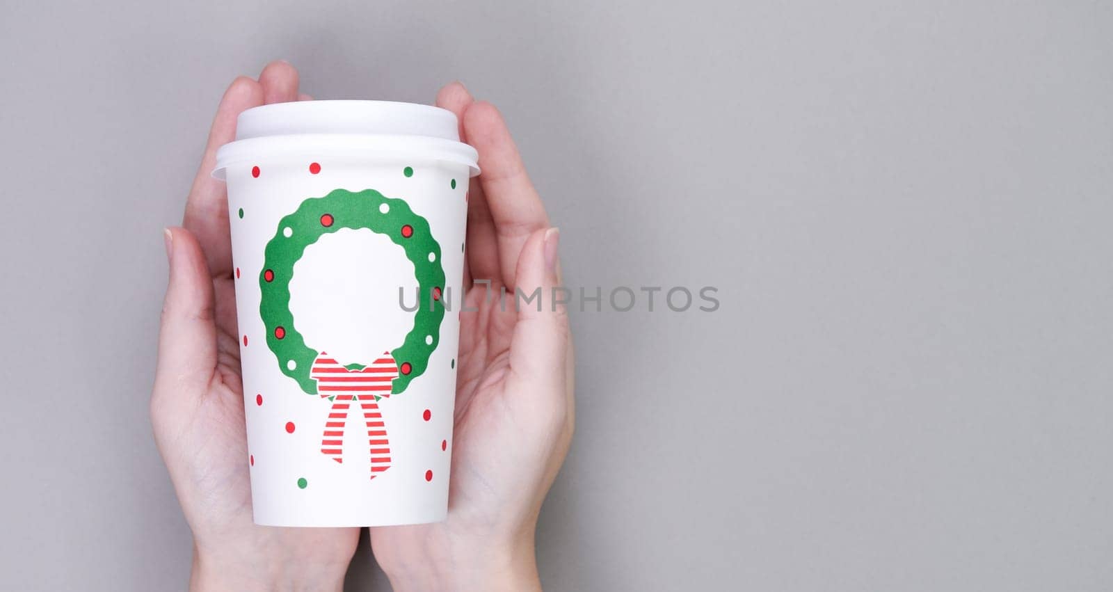 Hands holding craft paper coffee cup, grey background. Christmas or New Year holiday greeting paper coffee cup template. Flat lay, top view, copy space
