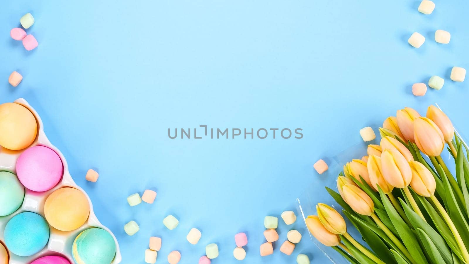 Spring flat lay with fresh yellow tulips colorful eggs on blue background. Easter, Mother day, birthday or Anniversary with copy-space. Sweet rainbow color Easter eggs, small pastel marshmallows