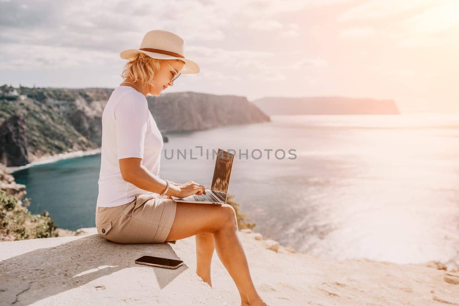 Digital nomad, Business woman working on laptop by the sea. Pretty lady typing on computer by the sea at sunset, makes a business transaction online from a distance. Freelance, remote work on vacation by panophotograph