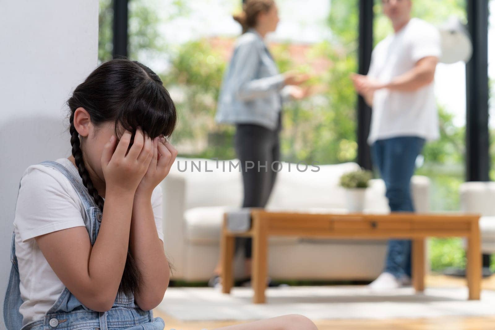 Stressed and unhappy young girl huddle in corner, cover her face while her parent arguing in background. Domestic violence at home and traumatic childhood develop to depression. Synchronos