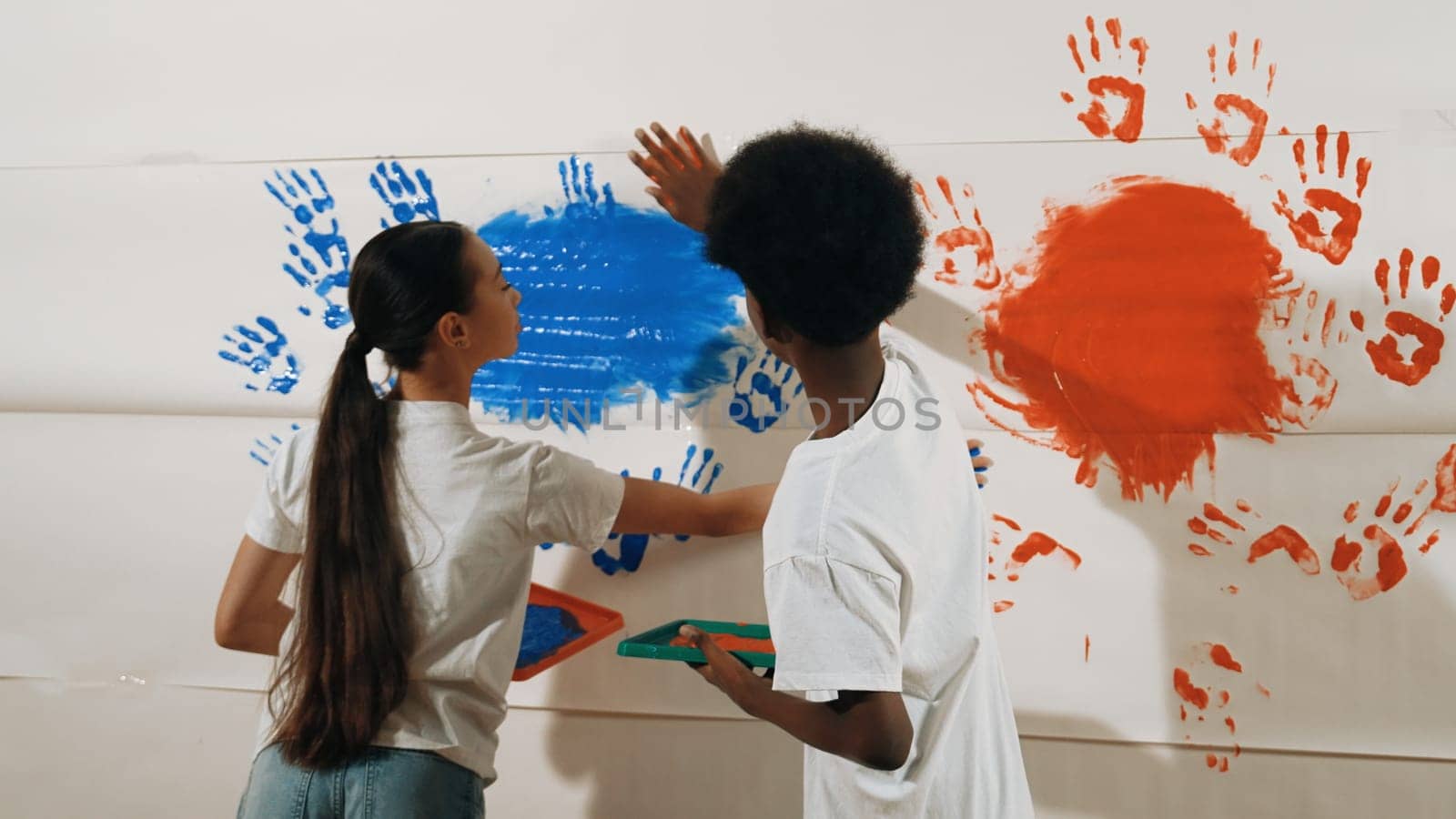 Diverse happy highschool girl and african boy paint the wall with water color in red and blue or contrast color by using their hands in art lesson. Colorful. Creative activity concept. Edification.