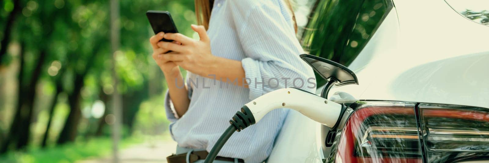 Young woman using smartphone online banking application to pay for electric car battery charging from EV charging station during vacation road trip at national park or summer forest. Panorama Exalt