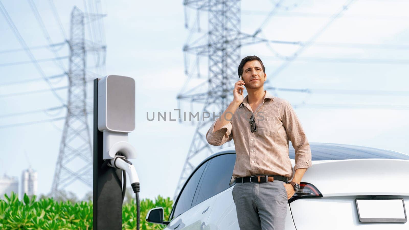 Man talking on the phone while recharge EV car battery at charging station connected to power grid tower electrical as electrical industry for eco friendly car utilization. Panorama Expedient